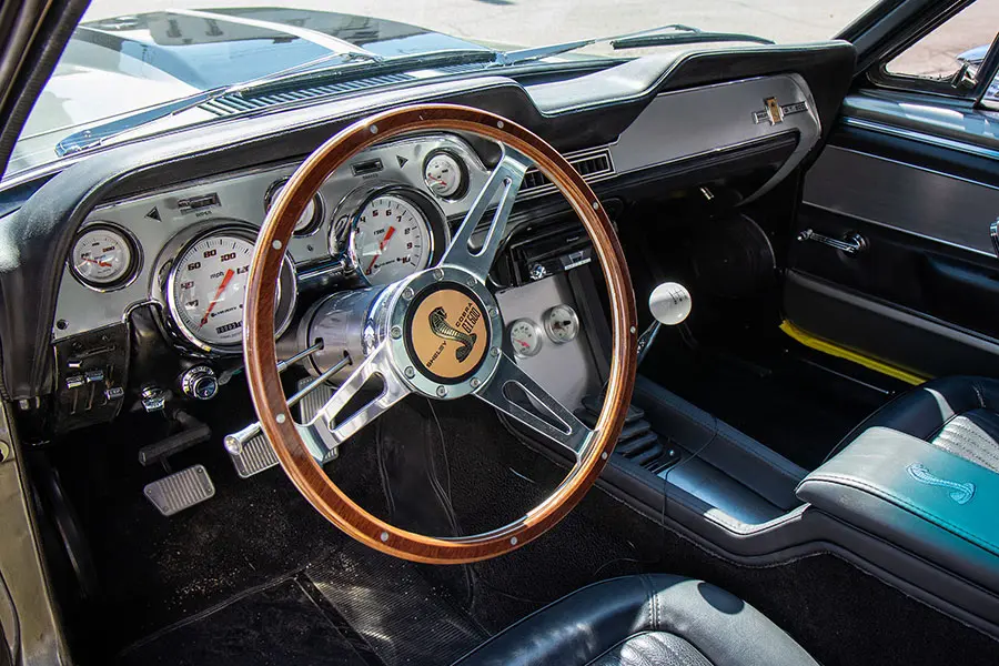 A restored Shelby Mustang interior with a wood-rim steering wheel and chrome gauges